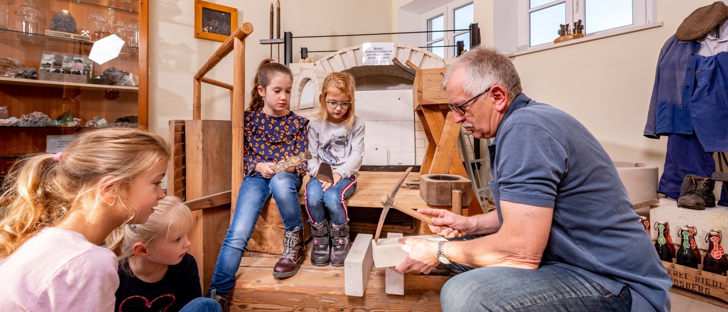 Mann zeigt vier Kindern Bearbeitung von Schamottesteinen in der Glasofenbauabteilung im Museum Plößberg