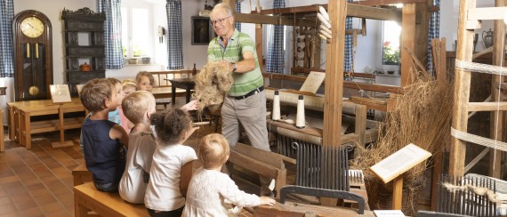Aufnahme in der Dauerausstellung mit historischen Webstühlen. Ein Mann gibt Flachsfasern an fünf Kinder.