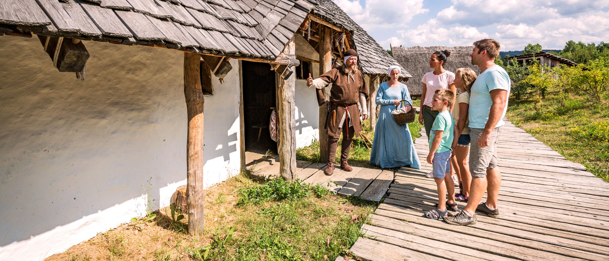 Mann und Frau in mittelalterlicher Kleidung zeigen einer Familie ein slawisches Langhaus